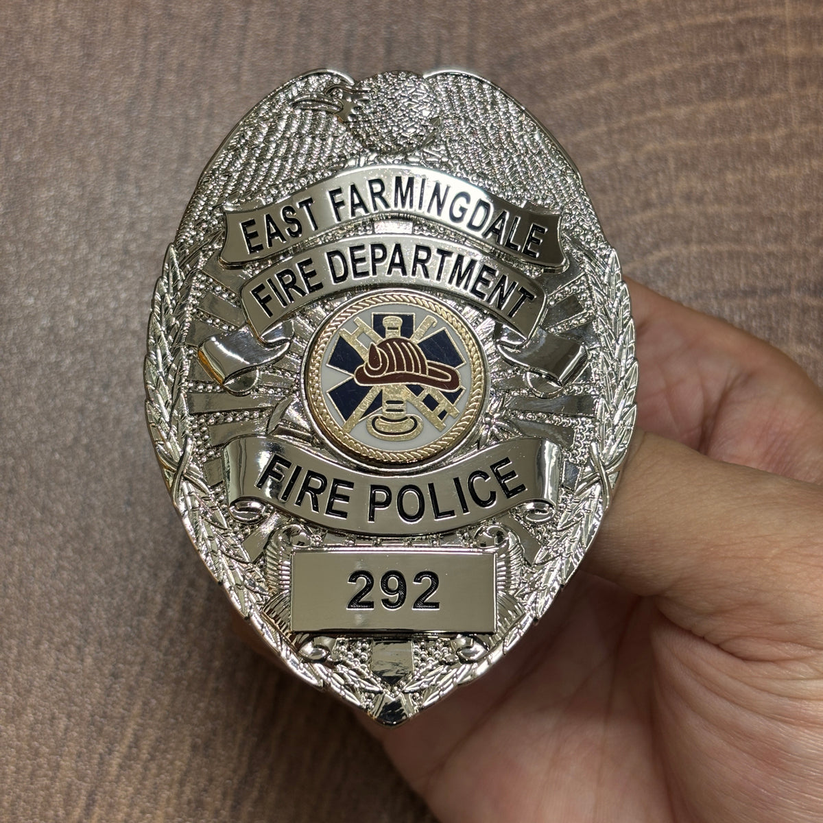Silver-tone oval fire department badge with black East Farmingdale lettering, firefighter helmet and crossed tools center emblem, FIRE POLICE role banner, and number 292
