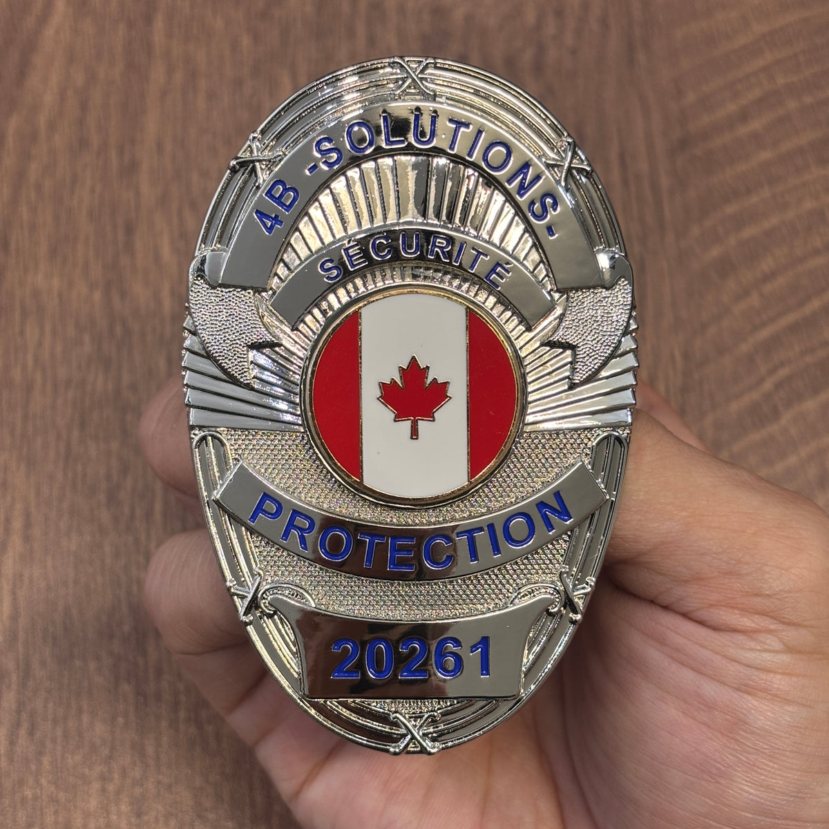 Silver-tone oval security badge with blue company lettering, French SÉCURITÉ text, Canadian maple leaf center emblem, PROTECTION banner, and number 20261