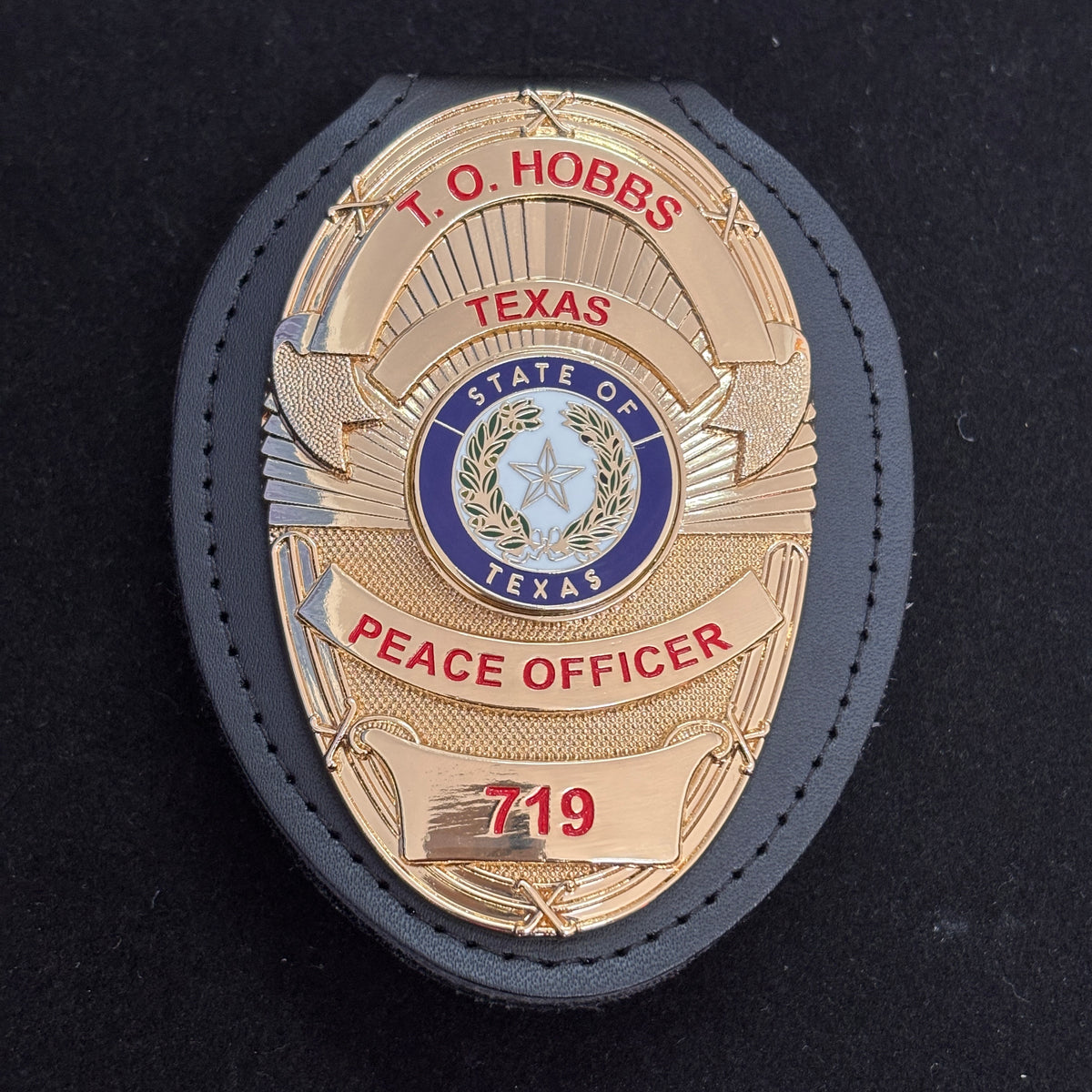 Two-tone gold-and-silver peace officer-style badge with red lettering, Texas-themed center emblem, number panel, and fitted black leather badge holder