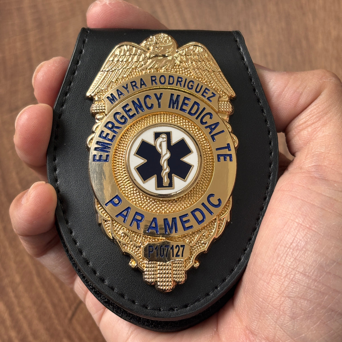 Gold-tone oval EMS badge in a black leather holder with eagle crest, blue lettering, Star of Life center emblem, personalized name panel, paramedic title, and ID number