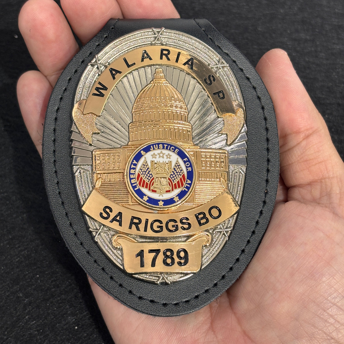 Two-tone personalized oval custom badge with architectural center design, colored emblem, black lettering, number panel, and fitted black leather holder