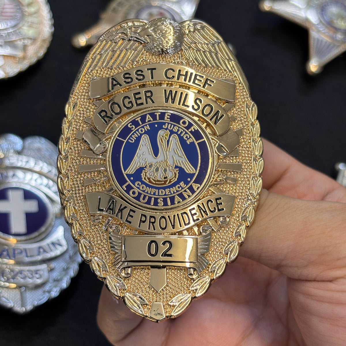 Gold-tone police-style custom metal badge with eagle crest, blue center emblem, curved title panels, personalized lower banner, and number panel