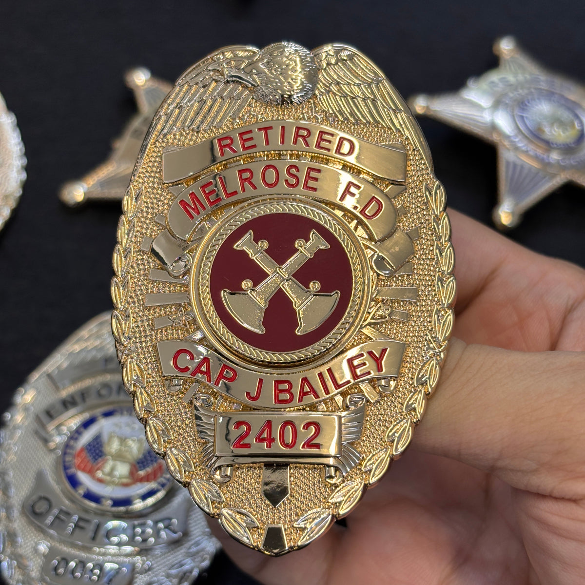 Gold-tone oval retired fire department badge with eagle crest, red RETIRED and MELROSE F.D. lettering, crossed fire tools, CAP J BAILEY name panel, and number 2402