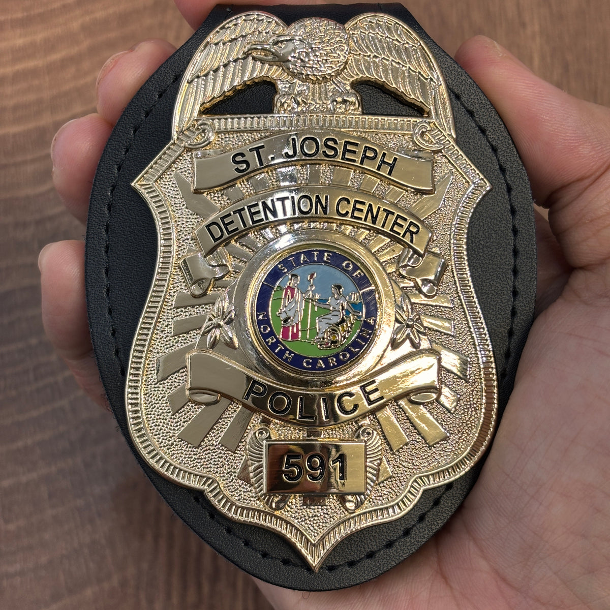 Two-tone shield badge in a black leather holder with eagle crest, ST. JOSEPH and DETENTION CENTER banners, North Carolina state emblem, POLICE title, and number 591
