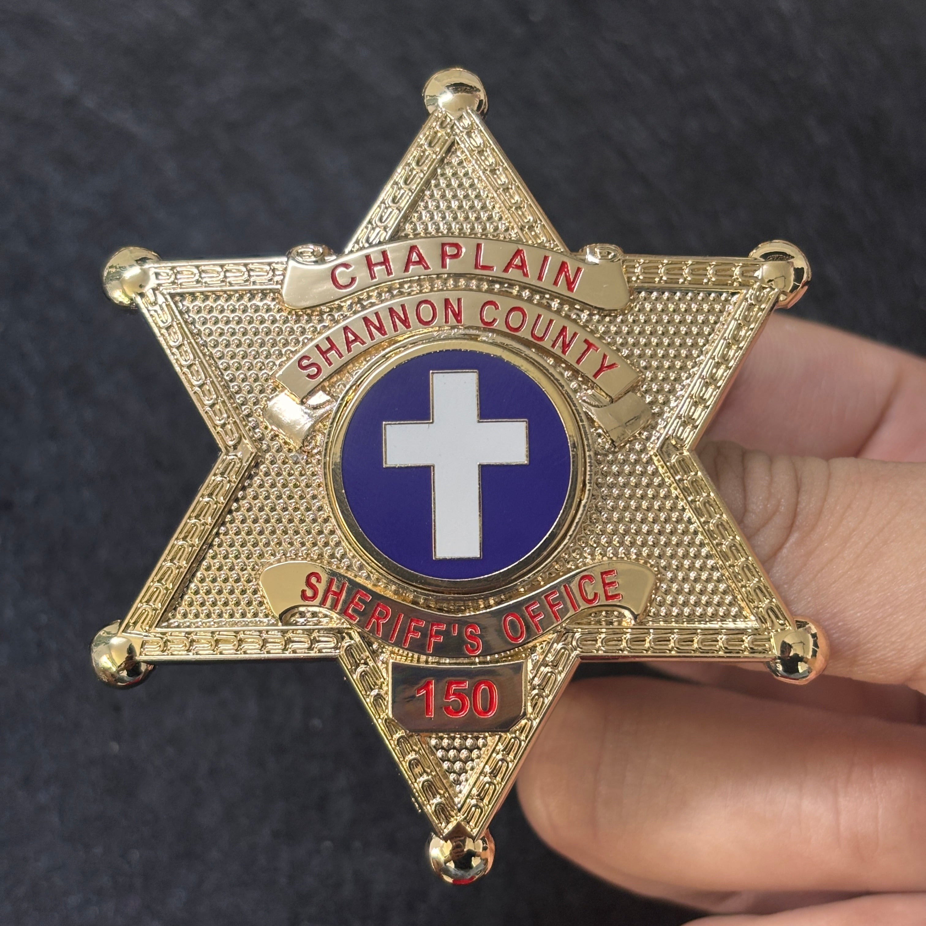 Gold-tone six-point custom chaplain badge with red lettering, a blue-and-white cross emblem, and identification number 150