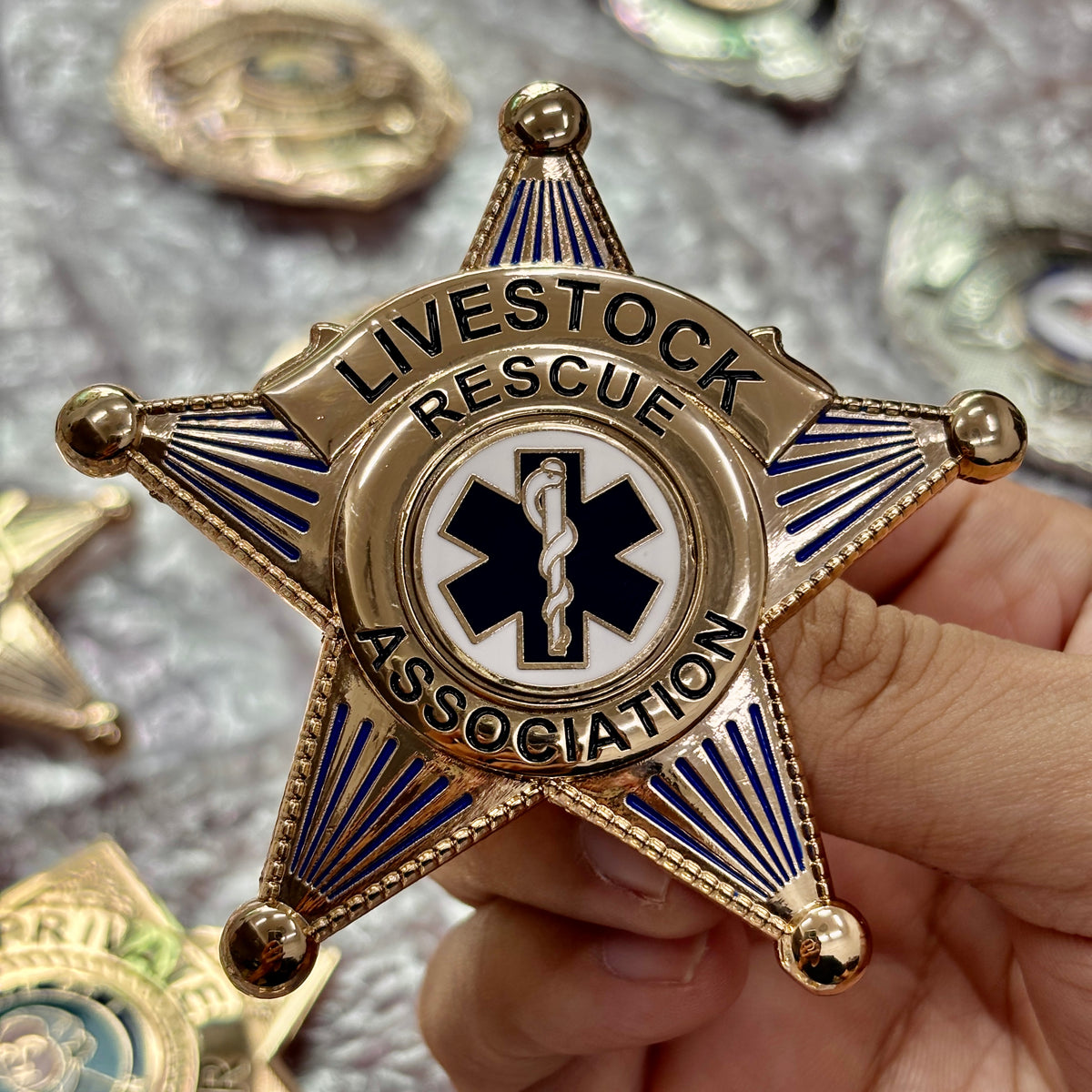 Gold-tone five-point LIVESTOCK RESCUE ASSOCIATION star badge with blue rays and a blue-and-white six-point medical-style center symbol