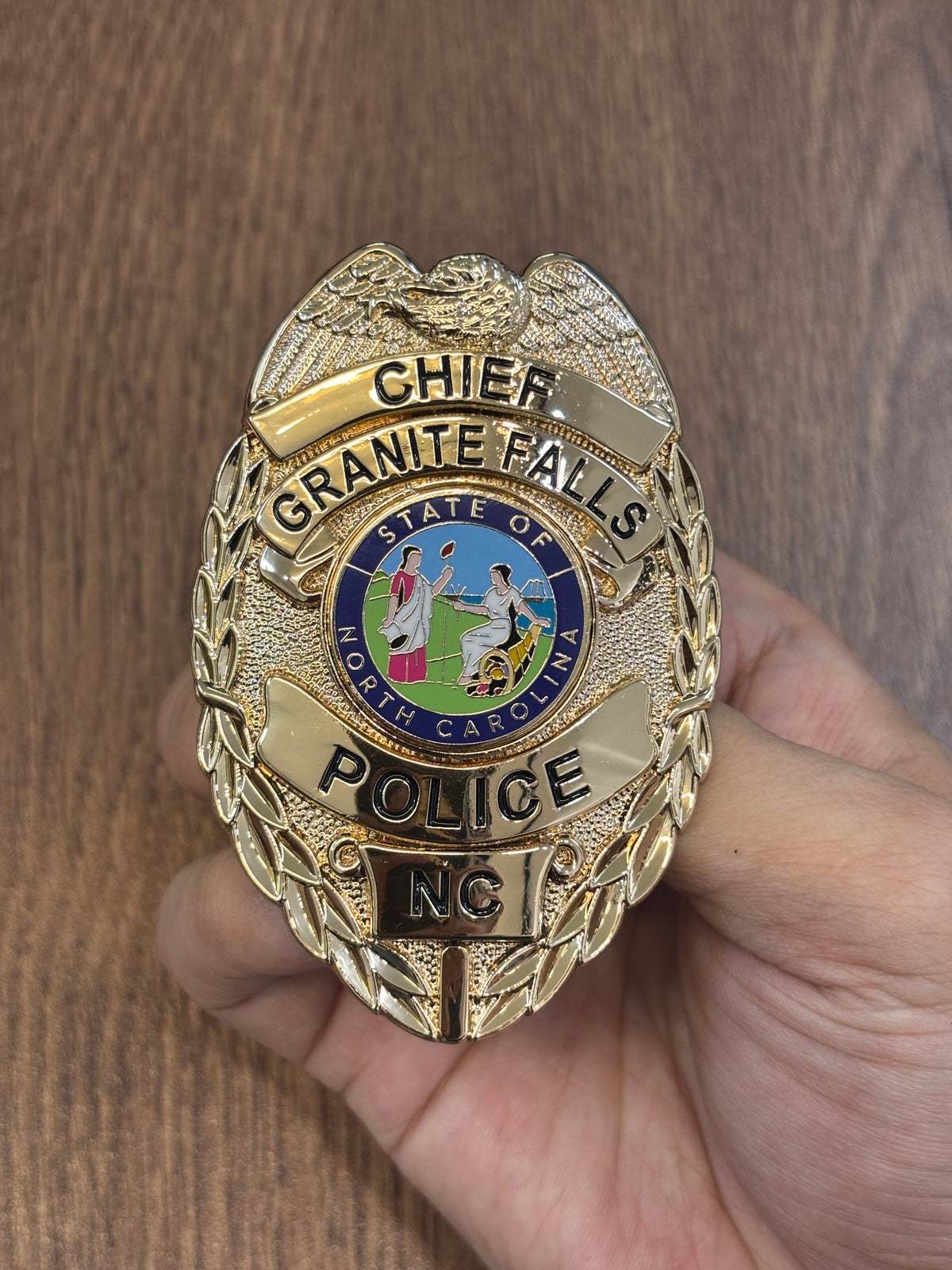 Gold-tone oval police officer-style custom badge with eagle crest, black lettering, North Carolina state emblem, agency name, rank panel, and lower location panel
